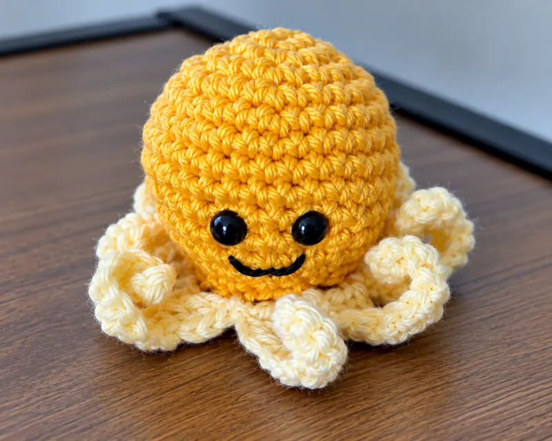 A crocheted toy octopus with a mustard-yellow head, cream tentacles and embroidered eyes, standing on a wooden surface.