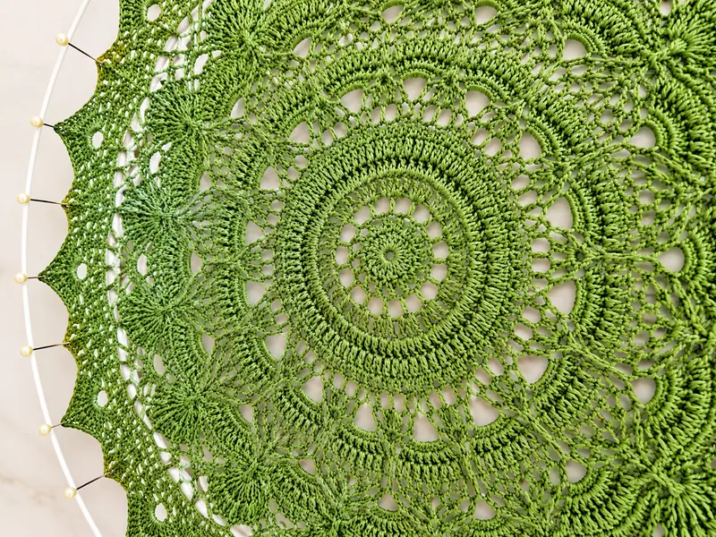 A green crocheted mandala doily with a scalloped edge, pinned out on a blocking hoop.