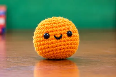 A small crocheted tangerine charm with an embroidered face, photographed against a plain green background.