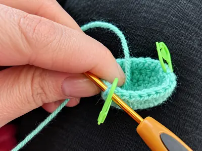 Close-up of a hand working a mint-green crochet bowl with an orange hook and green stitch markers.