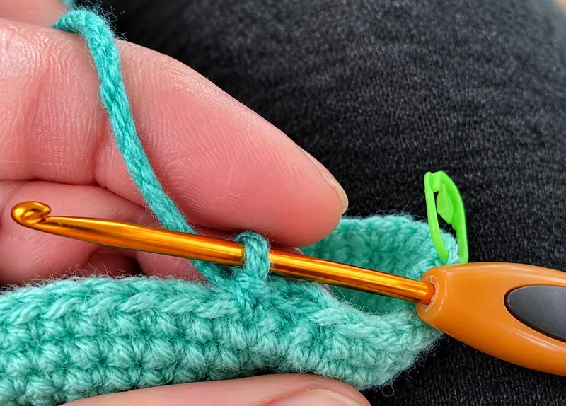 Close-up of a teal crocheted round in progress on an orange hook, with green stitch markers holding the working loop in place.